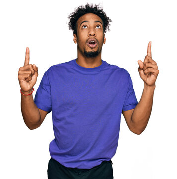 Young african american man with beard wearing casual purple t shirt amazed and surprised looking up and pointing with fingers and raised arms.
