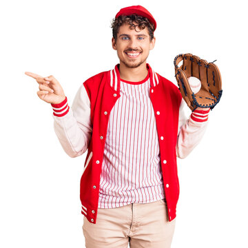 Young handsome man with curly hair wearing baseball uniform holding golve and ball smiling happy pointing with hand and finger to the side