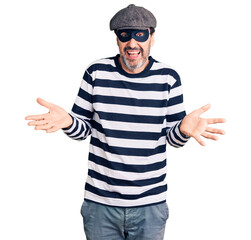 Naklejka premium Middle age handsome man wearing burglar mask clueless and confused expression with arms and hands raised. doubt concept.