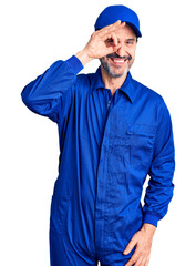Middle age handsome man wearing mechanic uniform doing ok gesture with hand smiling, eye looking through fingers with happy face.