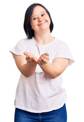 Brunette woman with down syndrome wearing casual white tshirt smiling with hands palms together receiving or giving gesture. hold and protection