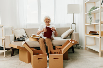 elderly woman sits on a sofa at home with boxes. collecting things with memories and moving and...