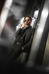 Serious man in classic black suit and sunglasses stand on the iron wall background and smoking a...
