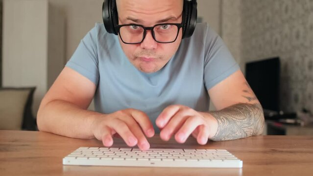 Geek funny man programmer looking at camera and typing on keyboard, listening music that plays in his headphones. Caucasian male nerd in glasses with tattooed hands sitting at desk in home and working