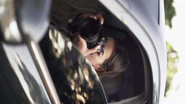 Side view of a female private detective sitting inside car photographing with slr camera.