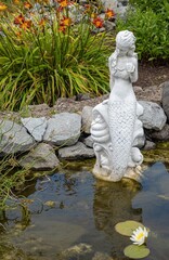 Fototapeta na wymiar A tranquil stone statue sculpture of a mermaid overlooks a still pond with day piles and a white lotus