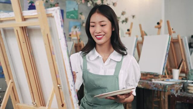 Woman paints picture on canvas with oil paints in her studio. Female artist painting picture in workshop. Young female artist painting on a canvas with a paintbrush and holding a color palette at home