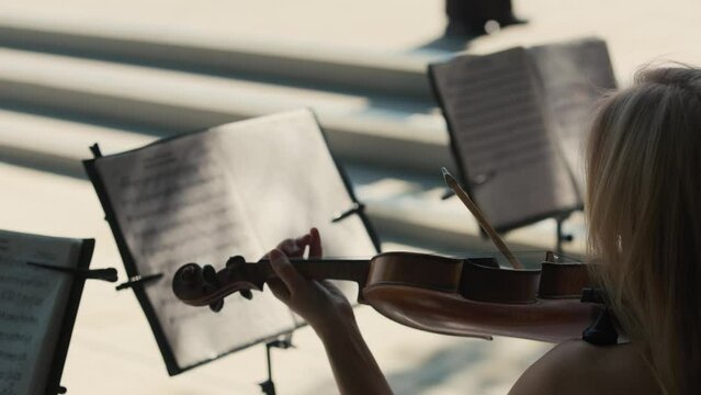 Close-up of the female musician sitting in a bright concert hall in front of a music stand with notes and playing the violin in the orchestra. Back view. The art of music.