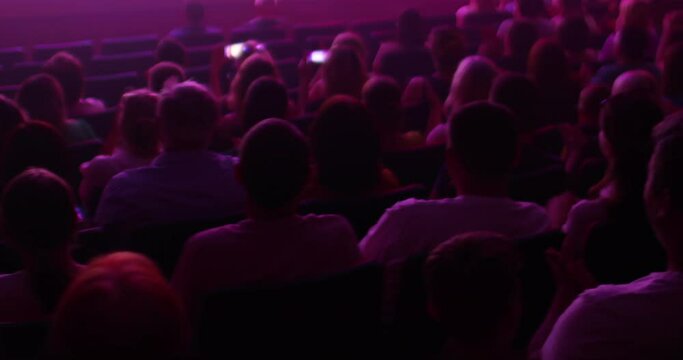 Silhouettes of spectators in the auditorium, watching the performance on stage and applauding. There is a dim violet light in the hall. Back view. Holding holidays, events, performances concept.