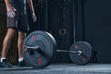 Male athlete stands back to the camera, looks down at the barbell on the floor in the gym closeup...