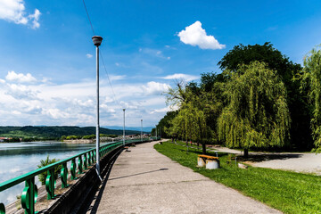 Fototapeta premium The alley in Targu Jiu central park next the Jiu river and in the background is the old bridge over the Jiu, also called Ferdinand's bridge. Targu jiu, Romania.