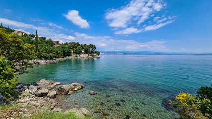 A panoramic view of the shore along Lovran, Croatia. There is a small town located between the lush trees. The Mediterranean Sea has turquoise color. Big boulders in the water. Sunny day. Calmness