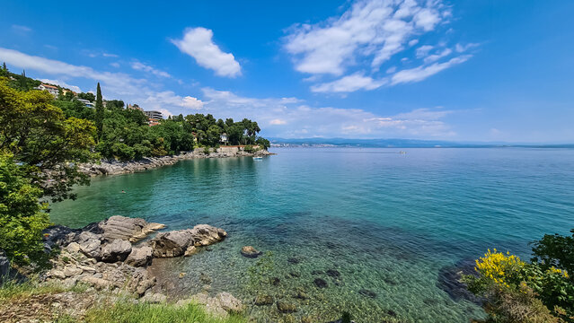 A panoramic view of the shore along Lovran, Croatia. There is a small town located between the lush trees. The Mediterranean Sea has turquoise color. Big boulders in the water. Sunny day. Calmness