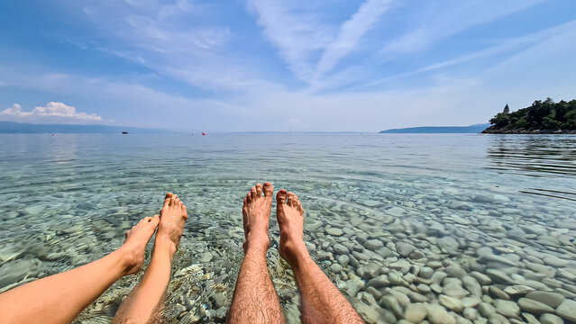 Two pairs of legs floating on the surface of Mediterranean Sea in Croatia. The sea is clear, with white stones on the bottom. An island visible in the back. Taking a break. Sunny and warm day.