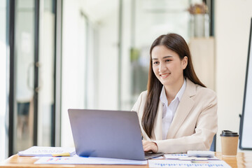 Asian female entrepreneur working in finance at home office analyzing financial graphs on documents with laptop on table in office