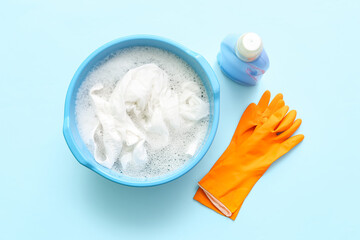 Plastic basin with dirty clothes, water, foam and orange rubber gloves on blue background