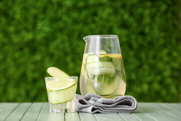 Glass and jug of lemonade with cucumber on green wooden table outdoors