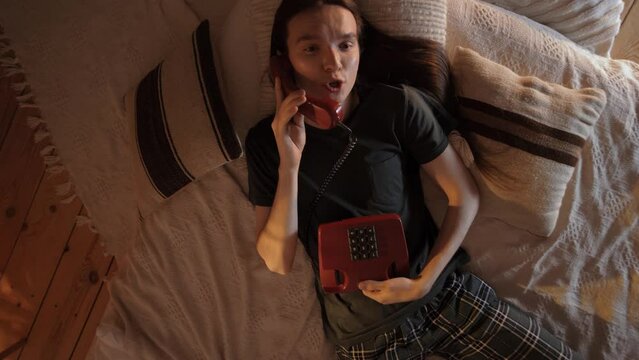 Attractive young guy with long hair lying in on a soft floor in a cozy room at night and talking on his home phone 