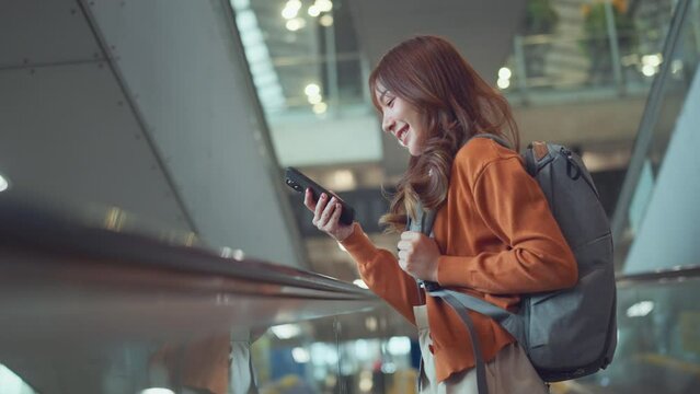 Asian tourist woman traveller with backpack using mobile phone while stand on moving walkway in airport terminal, Tourist journey trip concept.