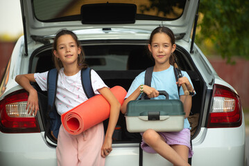 Two happy twin sisters with backpacks with a tent in their hands put tourist things in the trunk of a white car for going outdoors for recreation