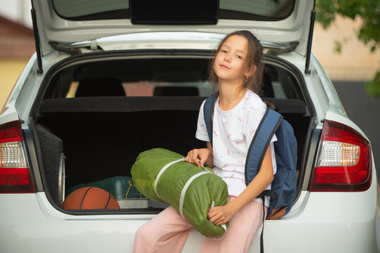 a happy girl in a white T-shirt with a backpack with a tent in her hands puts in the trunk of a white car for going outdoors for recreation