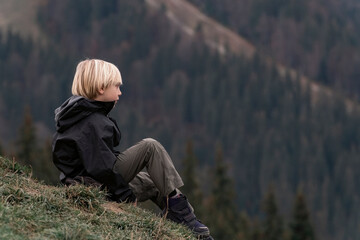 Naklejka na ściany i meble Blond boy in trekking clothes on hike during break. Child is relax on the mountainside. Boy sitting on hill