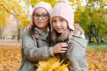 Two twin sisters posing for the camera with a bouquet of yellow leaves in the autumn park