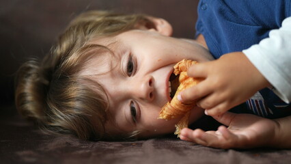 Child eating croissant one little boy lying on sofa eats carb food