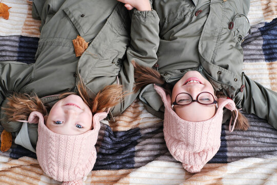 Two happy twin sisters lie on a blanket in an autumn park. The girls smile and look at the camera.