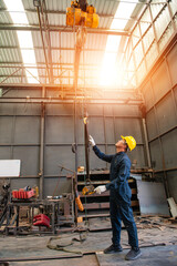 engineer man wearing bodysuit and yellow contruction helmet holding his arms with white gloves checking a hook of lifting hoist in lathe factory background with light and flare