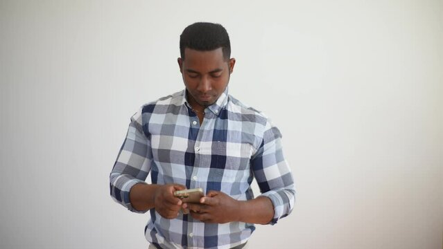 Studio portrait of laughing african american man in plaid shirt using holding mobile cell phone chatting standing on white isolated background, looking to device screen with cheerful expression.