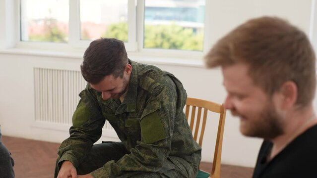 Group of cheerful male veterans in camouflage uniform talking and laughing sitting in circle during PTSD group therapy session, slow motion. Concept of mental health, psychotherapy, social issues.