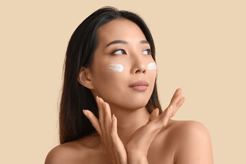 Beautiful young Asian woman with cream on her face against beige background