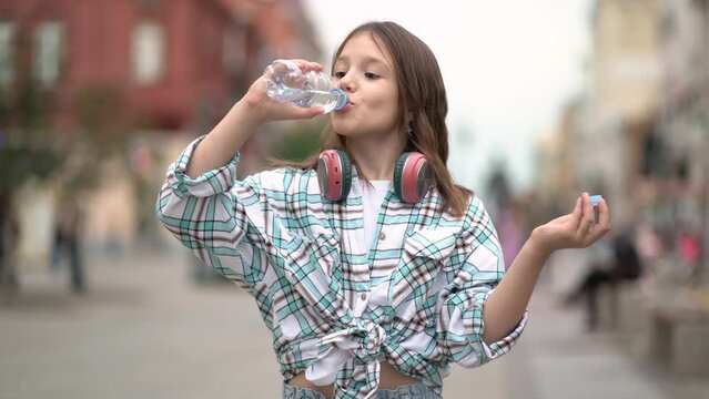 Girl quenches thirst drinks cool water from a plastic bottle on the street in a shirt and headphones.