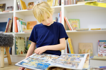 A preteen boy leafing through a book while sitting at the bookshelves at home, in a school library...