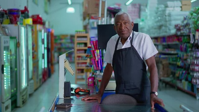 One black senior employee of Supermarket standing in cashier checkout portrait wearing apron with neutral expression. Serious African American staff of Grocery Store