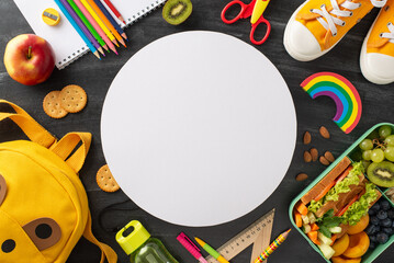 Nutritious school lunch idea. Top view of eco-friendly lunchbox filled with fresh, wholesome food, sport shoes, bag, vibrant stationery on chalkboard backdrop with circle for text or advertisement