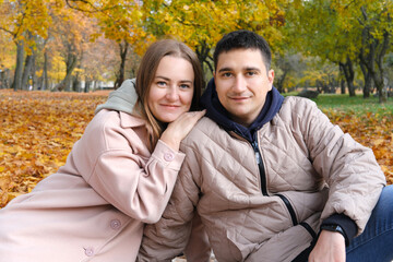 Young couple, husband and wife, sit on a blanket in an autumn park, smile and look at the camera