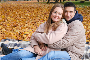 A young couple, husband and wife, are sitting on a blanket in an autumn park, the husband is hugging his wife, the couple is smiling and looking at the camera
