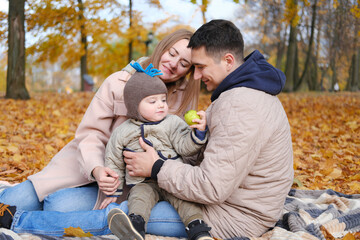 Family weekend. Young parents with a little son spend time together in the autumn park sitting on a blanket. Child eats an apple