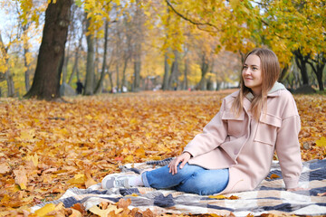 A beautiful young girl in jeans and a pink coat sits on a plaid blanket in an autumn park. The girl smiles and looks away
