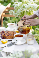 Summer elegant picnic. Woman in white dress purring tea from teapot at wooden table in garden. Beautiful dishwater, white porcelain, tablecloth, homemade cherry pie.
