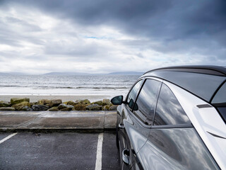 Fototapeta na wymiar Dark color car parked by the ocean with view on a stunning ocean landscape with mountains in the background. Dramatic cloudy sky. Galway bay, Ireland, Travel and sightseeing.