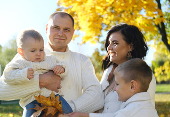 A happy family of four spend time in the autumn park. Mother and eldest son look at the youngest child, father smiles and looks at the camera