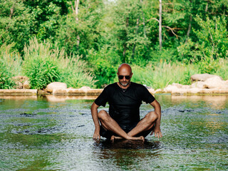 Bald male in dark wet shirt and shorts sitting in a stream and practice yoga relaxation. Selective focus. Green nature background. Healthy habit to battle stress and reach inner peace. Yoga guru.