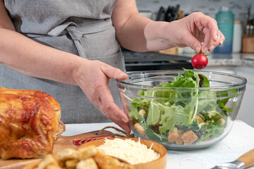 Cooking Caesar salad. Cook adds cherry tomatoes to glass bowl with salad in home kitchen.