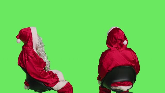 Santa claus posing on greenscreen background, young man dressed as saint nick sitting on chair and waiting. Acting impatient while he wears iconic festive costume with white beard.
