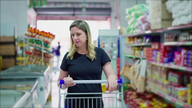 Female Consumer Walking In Supermarket Aisle Browsing Products On Shelf. One Caucasian Woman In 40s Pushing Shopping Cart