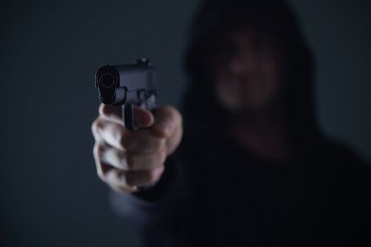 robber with gun aiming into camera. Man in hood threatens with firearm. Weapon in person's hands. Murderer or armed thief. Criminal with pistol.