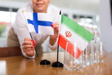 Little flag of Iran on table with bottles of water and flag of Finland put next to it by positive young woman in meeting room